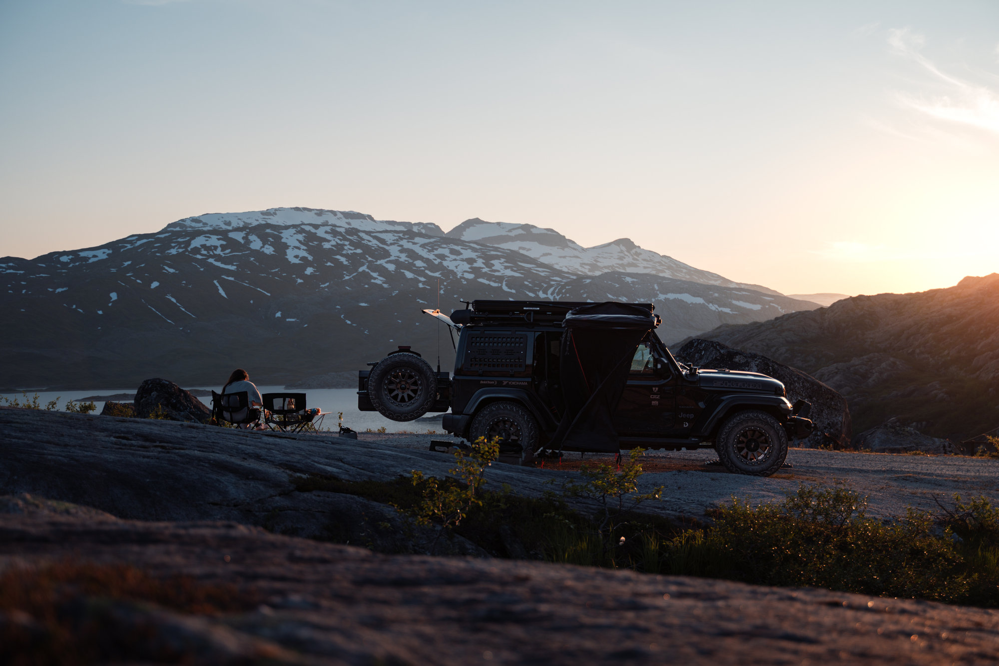 The Jeep parked on rock by a fjord at sunset, snow-capped mountains behind